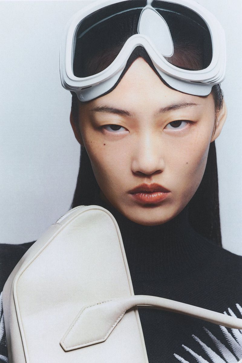 Portrait of an Asian woman wearing ski goggles and holding a white purse in front of a plain, light-colored background.