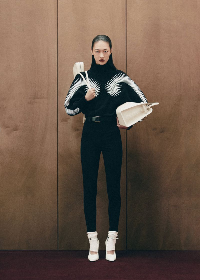 Fashion model in studio wearing black patterned sweater, black pants, and white heeled shoes holding a white handbag with wood background.