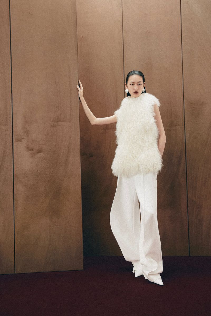Fashion shot: Woman poses in a white, sleeveless, shaggy faux-fur top and white wide-leg pants against a wood-panel wall indoors.