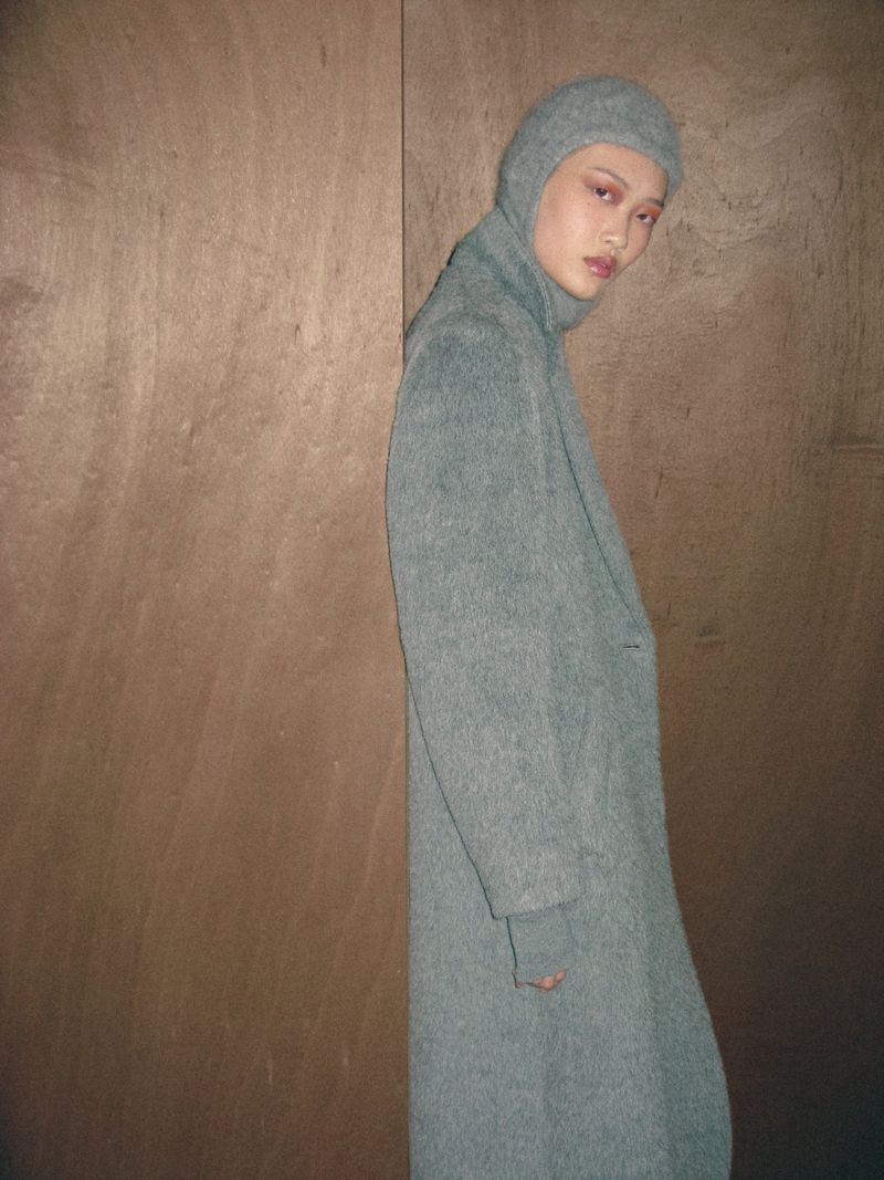 Model Ruiyi Zhang poses in a textured grey hooded wool coat against a plywood backdrop.