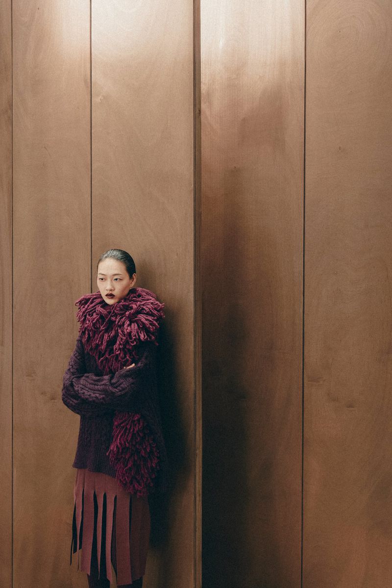Asian woman in a dark red sweater and fringed skirt standing against a wood-paneled wall in a studio setting with dramatic lighting.