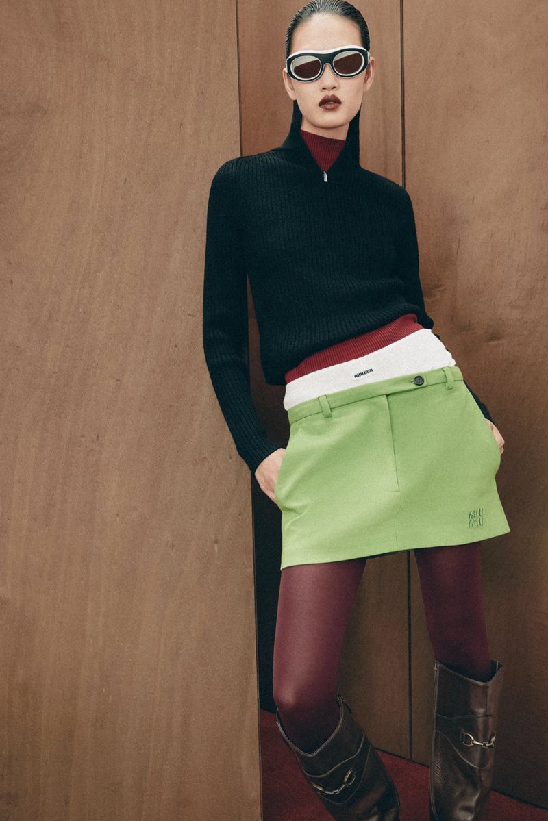 A stylish adult female poses indoors wearing sunglasses, a black top, green skirt, and brown tights and boots against a wood panel.