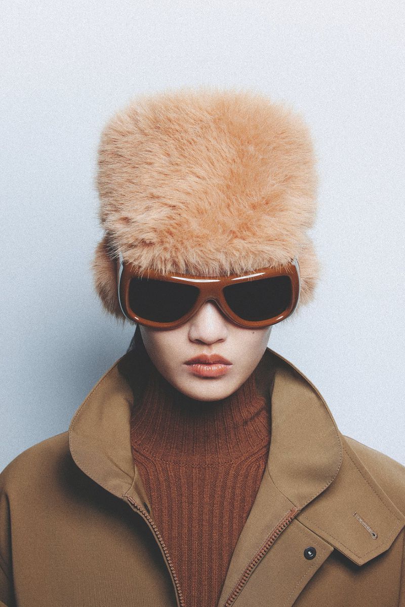 Woman in fur hat and oversize sunglasses, wearing a brown turtleneck and coat in a studio portrait against a white backdrop.