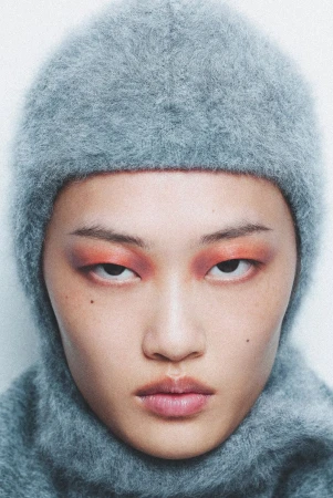 Close-up studio shot of a young adult female wearing a gray fuzzy balaclava and vibrant orange makeup around her eyes, looking at the camera.