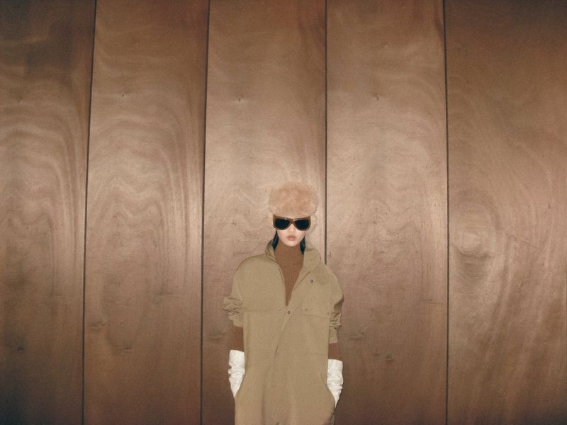 Woman in vintage fur hat and tan jumpsuit poses in front of a minimalist wood-paneled wall, wearing sunglasses and white gloves.