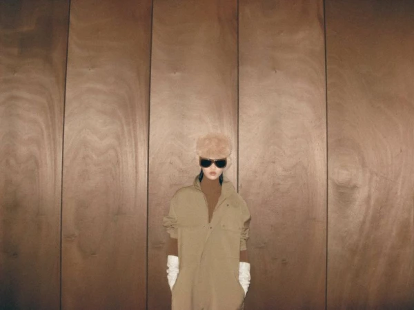 Woman in vintage fur hat and tan jumpsuit poses in front of a minimalist wood-paneled wall, wearing sunglasses and white gloves.