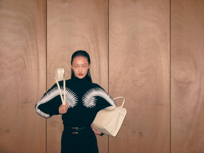 Asian woman posing with a bag, wearing a black turtleneck sweater with white detail, black pants with belt against wood paneling.