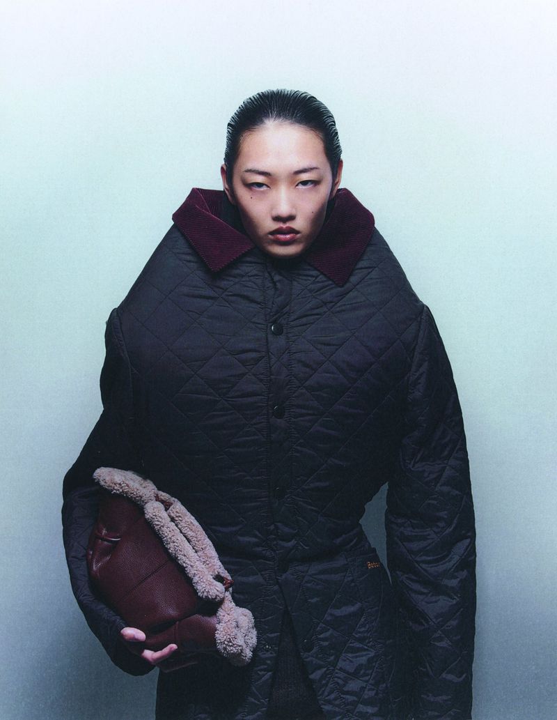 Asian female model in a black quilted coat holding a brown faux fur purse against a light green background in a studio shot.