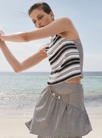 A young woman poses on a beach wearing a striped tank top and matching skirt, with the ocean and sky in the background on a sunny day.