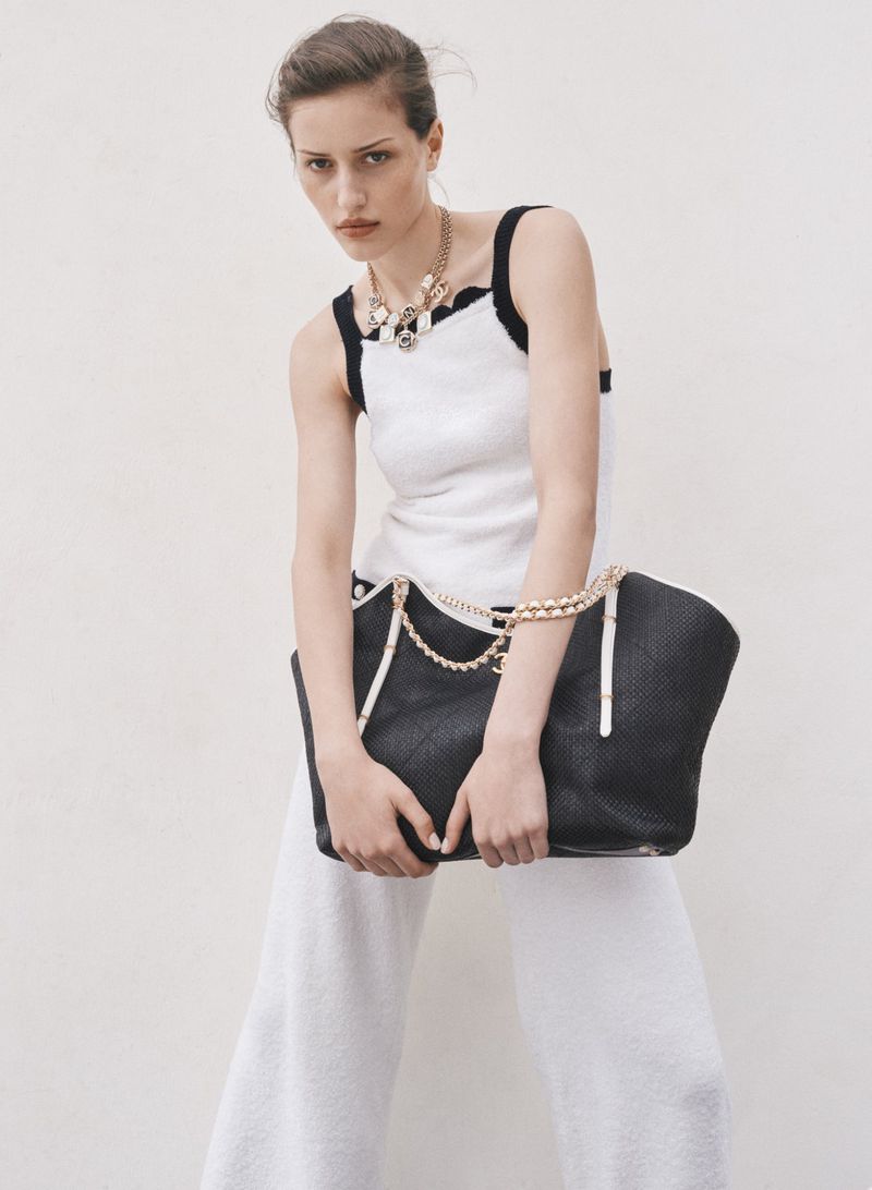 Model posing in a white tank top and wide-leg trousers while holding a large black tote bag against a white backdrop.