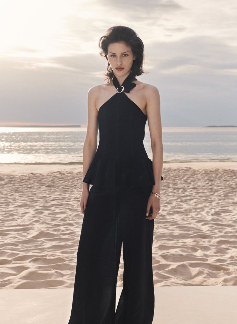 Elegant woman in a black halter top and wide-legged trousers poses on a sandy beach with a sea and sky backdrop at dusk.