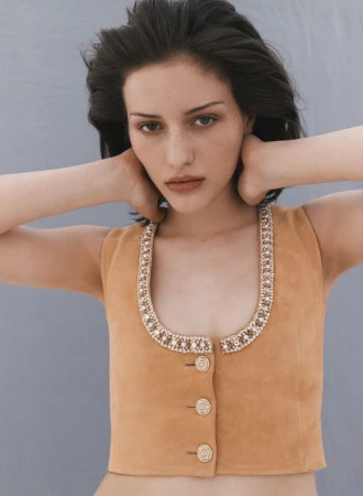 Brunette woman in a tan button-up vest with beaded trim and hands behind her head posing in front of a solid light blue backdrop.