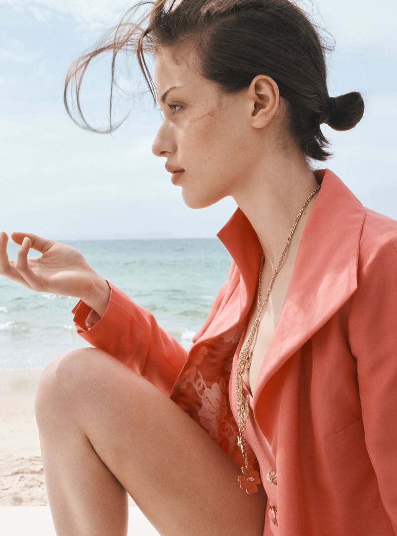 Young woman with stylish ponytail and coral blazer poses gracefully on a beach against a tranquil sea and sky backdrop on a sunny day.
