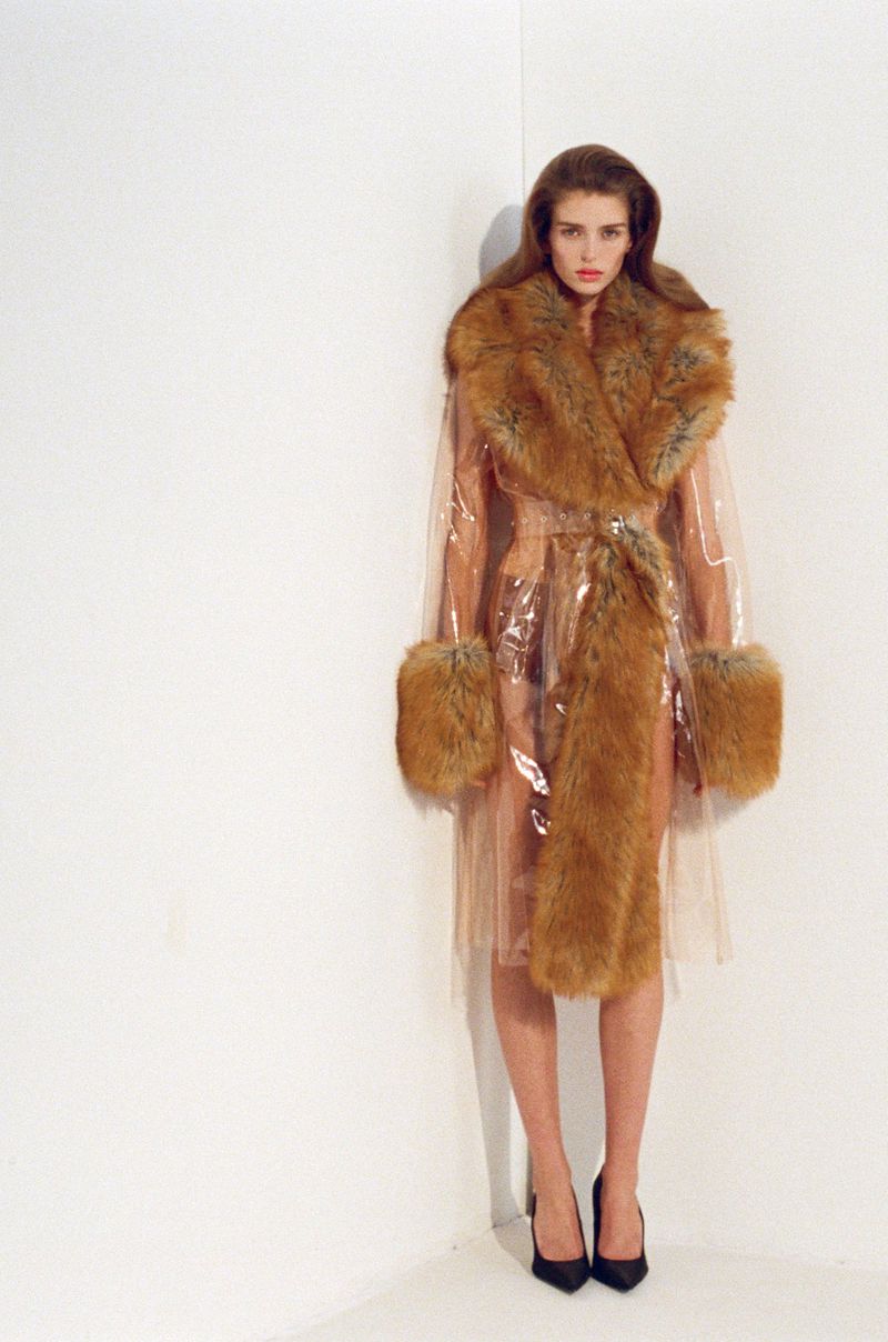 Woman in clear coat with fur trim and black heels poses in a photo studio against a white wall, looking at the camera.