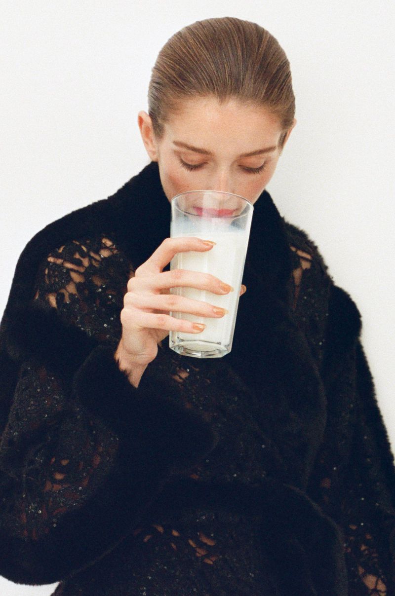 Woman in black lace robe drinks milk from glass against white background. Close-up beauty shot shows healthy lifestyle, studio lighting, and neutral colors.