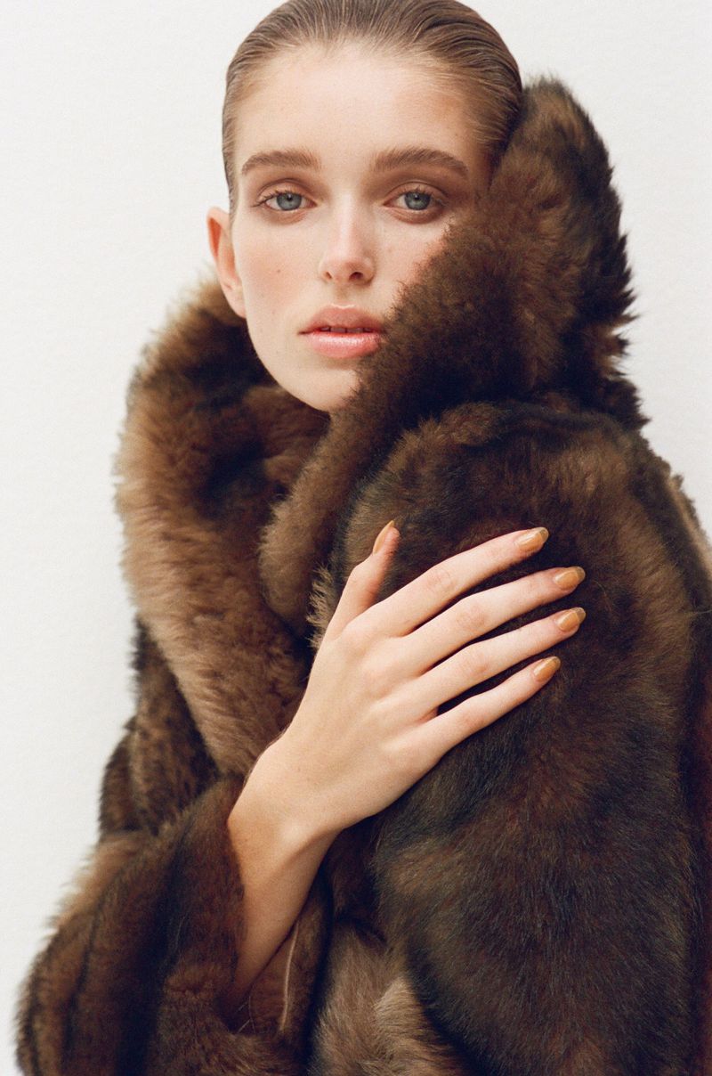 Close-up of a young adult woman wearing a brown fur coat, with a neutral expression against a clean, white studio backdrop.