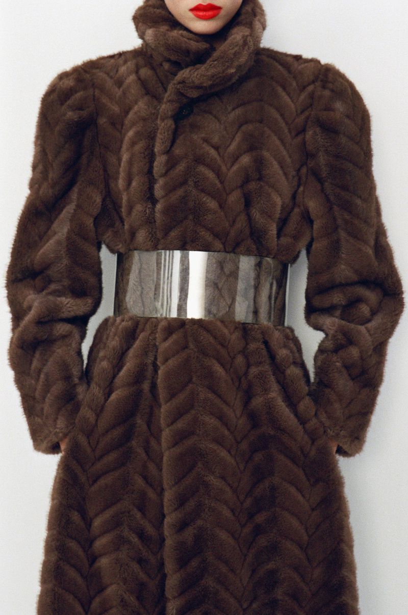 Elegant woman in a brown herringbone pattern mink coat with an oversized silver belt and bold red lipstick, against a plain white background.
