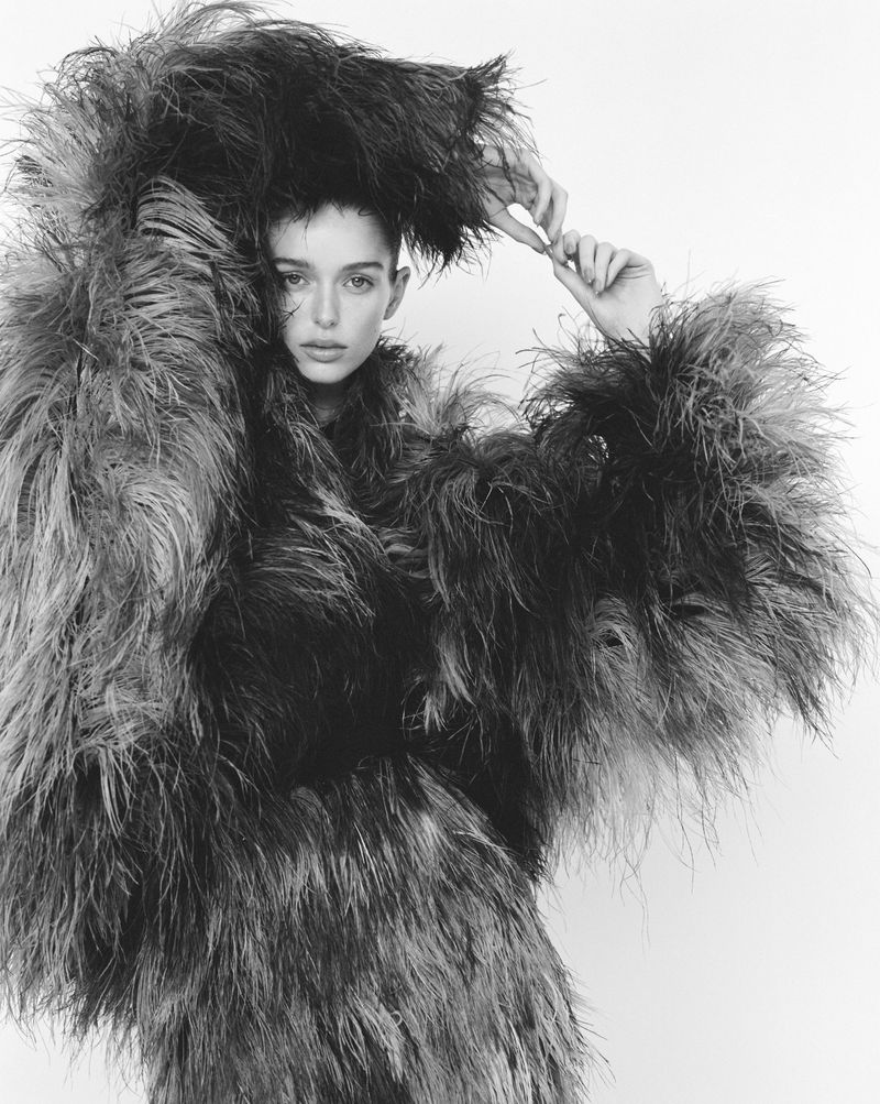Monochrome portrait of a young woman in a feather coat posing against a white backdrop, showcasing high fashion and editorial style.