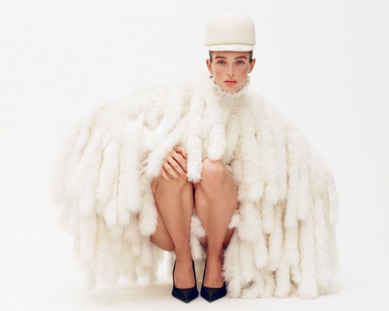 Fashionable adult female poses indoors in a crouched position wearing a white fluffy coat, black heels, and a white cap against a white backdrop.