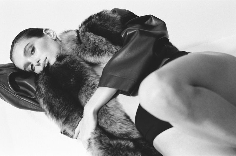 Model Stella Hanan reclining in a textured fur and leather jacket against a white studio background.