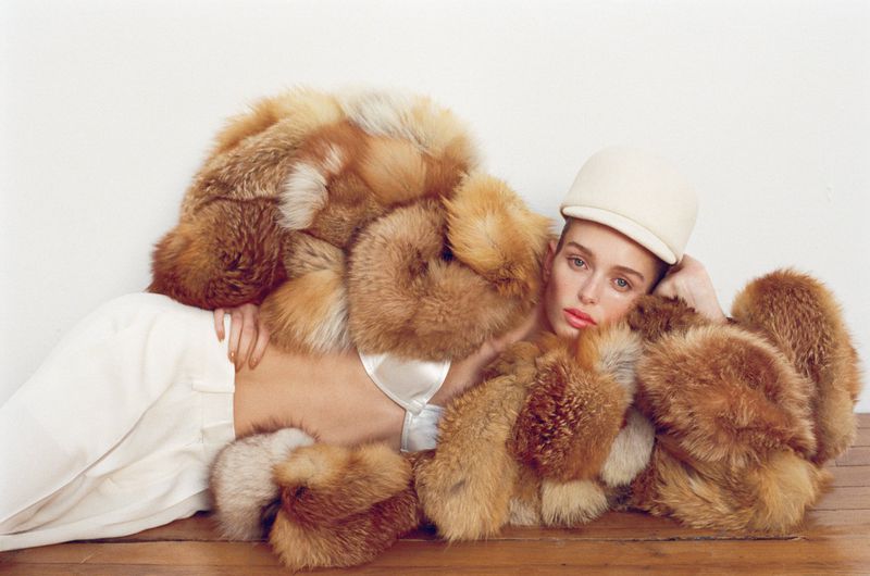 Fashion photo of a young adult woman in white pants and a matching cap lying down in an oversized fox fur coat on a wooden surface.