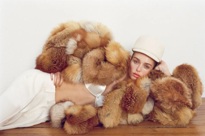 Fashion photo of a young adult woman in white pants and a matching cap lying down in an oversized fox fur coat on a wooden surface.