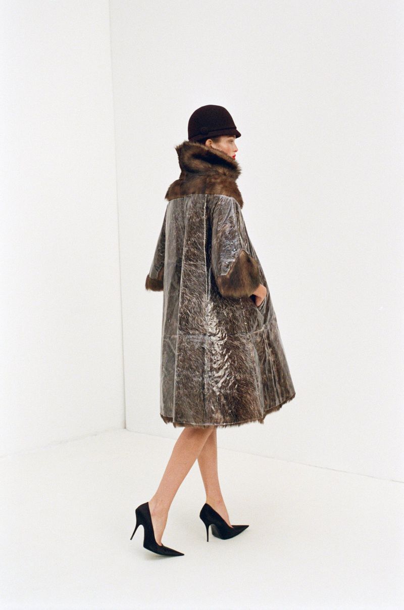 A woman wearing a fur-trimmed coat and hat is walking in a white studio. She is wearing black high heels. The photo is a fashion editorial.
