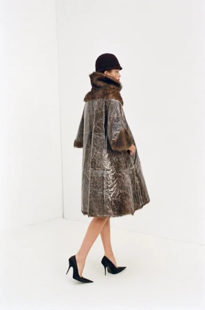 A woman wearing a fur-trimmed coat and hat is walking in a white studio. She is wearing black high heels. The photo is a fashion editorial.
