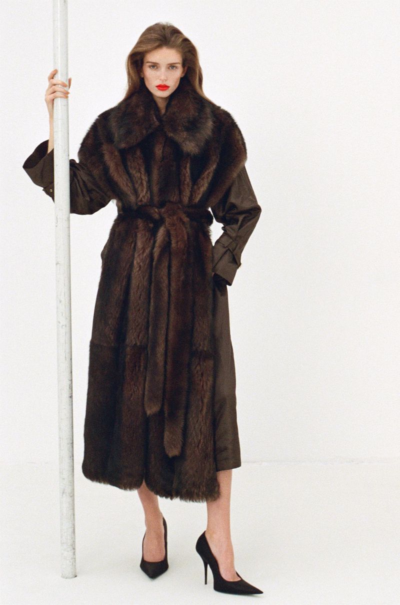 Fashion model in a brown fur coat and black heels poses in a studio, leaning against a white pole, creating a chic and editorial image.