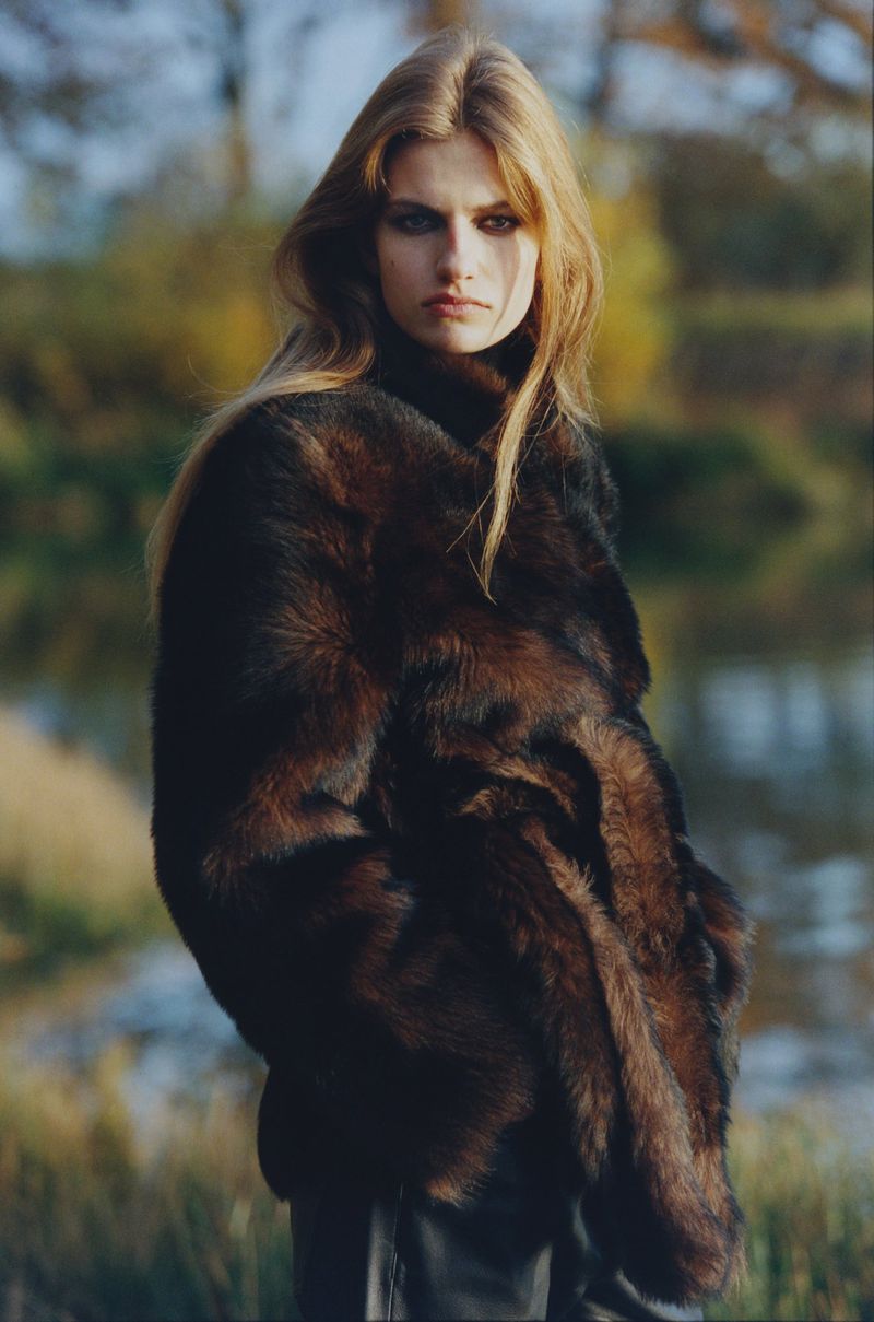 Blonde model in a brown fur coat stands outdoors near water and trees in a fashion shot on an overcast day.