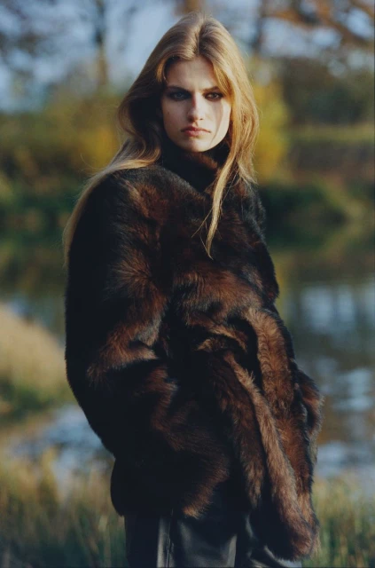 Blonde model in a brown fur coat stands outdoors near water and trees in a fashion shot on an overcast day.