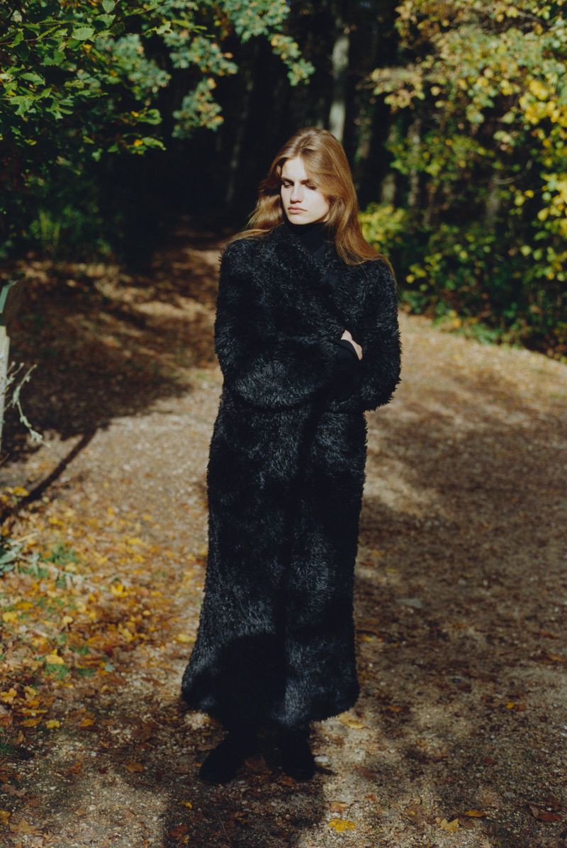 Model wearing a full-length textured black coat standing on a sunlit woodland trail.