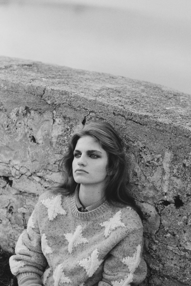 Portrait of a young woman with long wavy hair in a star-patterned sweater, leaning against a cracked stone wall outdoors in black and white.