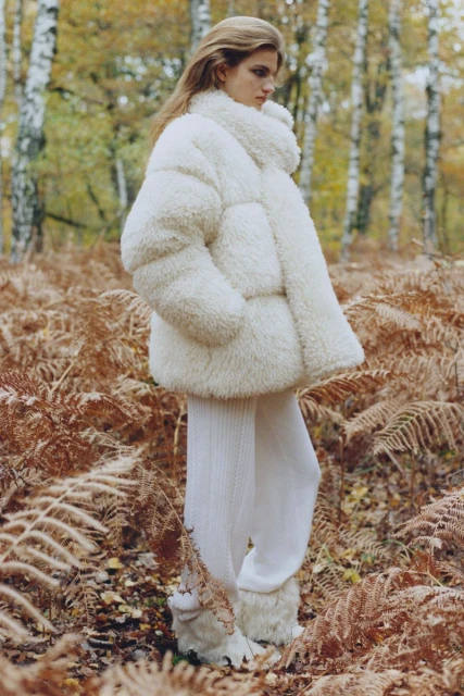 A woman with fair skin and blonde hair wears a fuzzy white coat and stands among brown plants and trees in an outdoor wooded setting.
