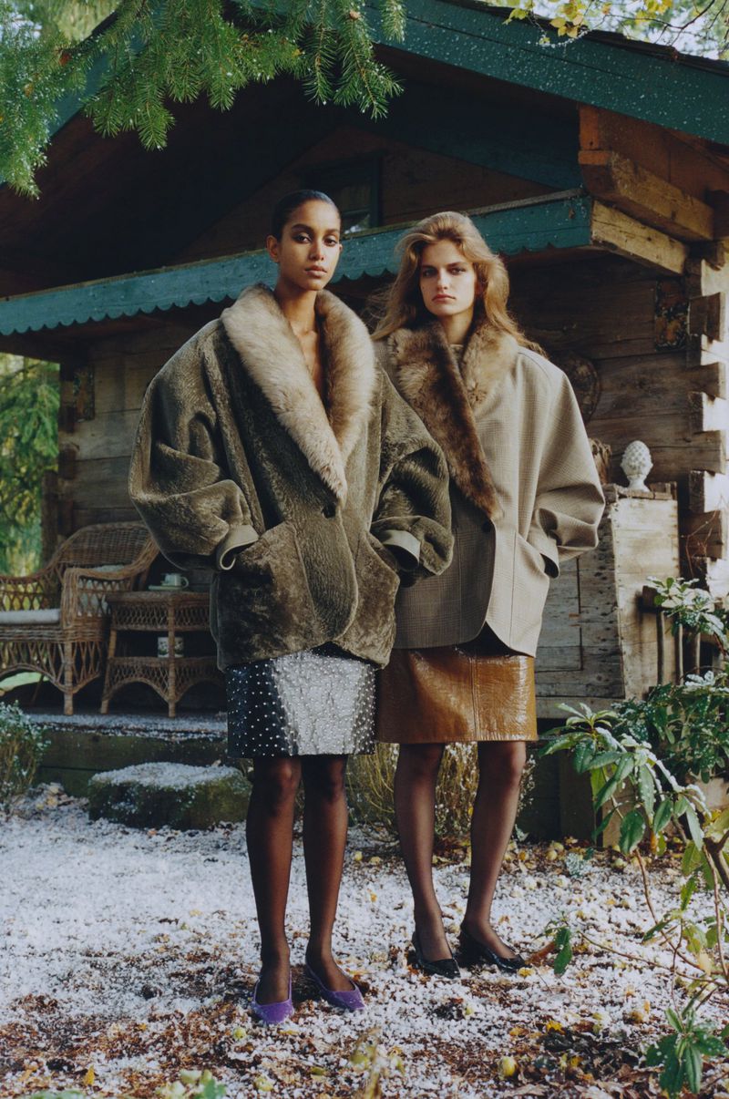Two women pose outdoors in oversized coats with fur collars, paired with mini skirts and stockings, against a log cabin backdrop.