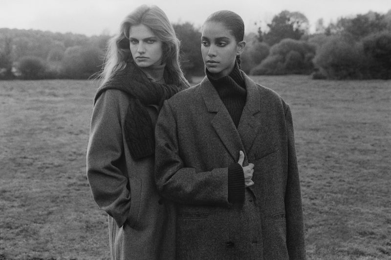 Two female models in oversized coats and turtlenecks posing outdoors in a grassy field under an overcast sky.