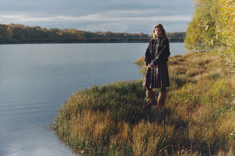 A woman in a tartan kilt and black sweater stands on a grassy shore, near a lake and forest under a cloudy sky in natural light.