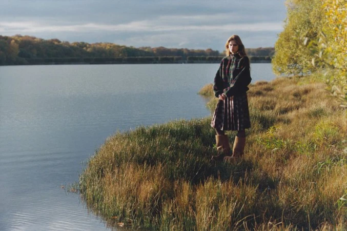 A woman in a tartan kilt and black sweater stands on a grassy shore, near a lake and forest under a cloudy sky in natural light.
