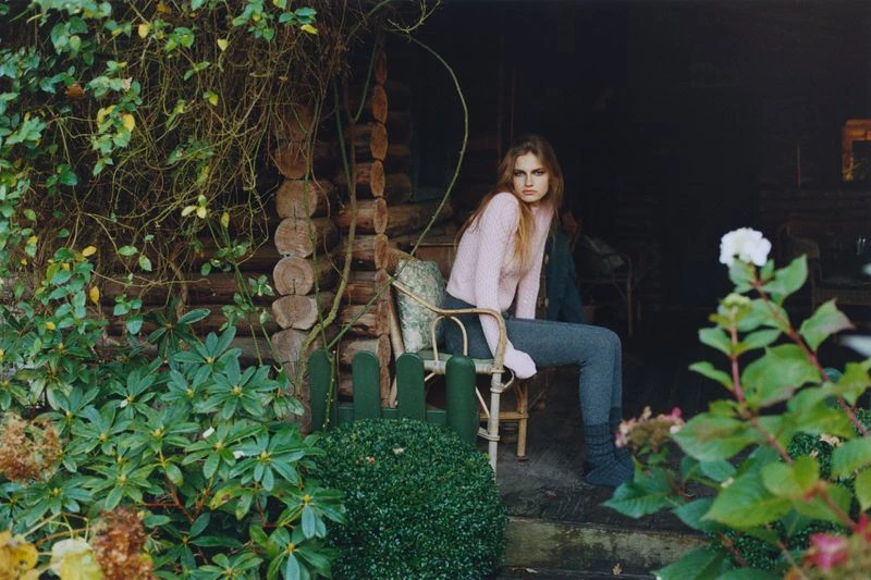 Woman in pink sweater and grey pants poses on a wooden chair on porch of log cabin surrounded by green plants and white flowers.