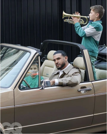 Man sits in convertible car as boy stands behind him playing trumpet. Another child is in the passenger seat in daytime. 