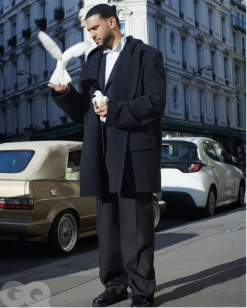 Stylish man in oversized coat poses with white doves in urban setting, featuring parked cars and building facade in daytime lighting.