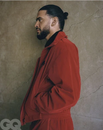 Man in red jacket and sweatpants poses in side profile with a stone wall background, showcasing a casual yet stylish fashion aesthetic.