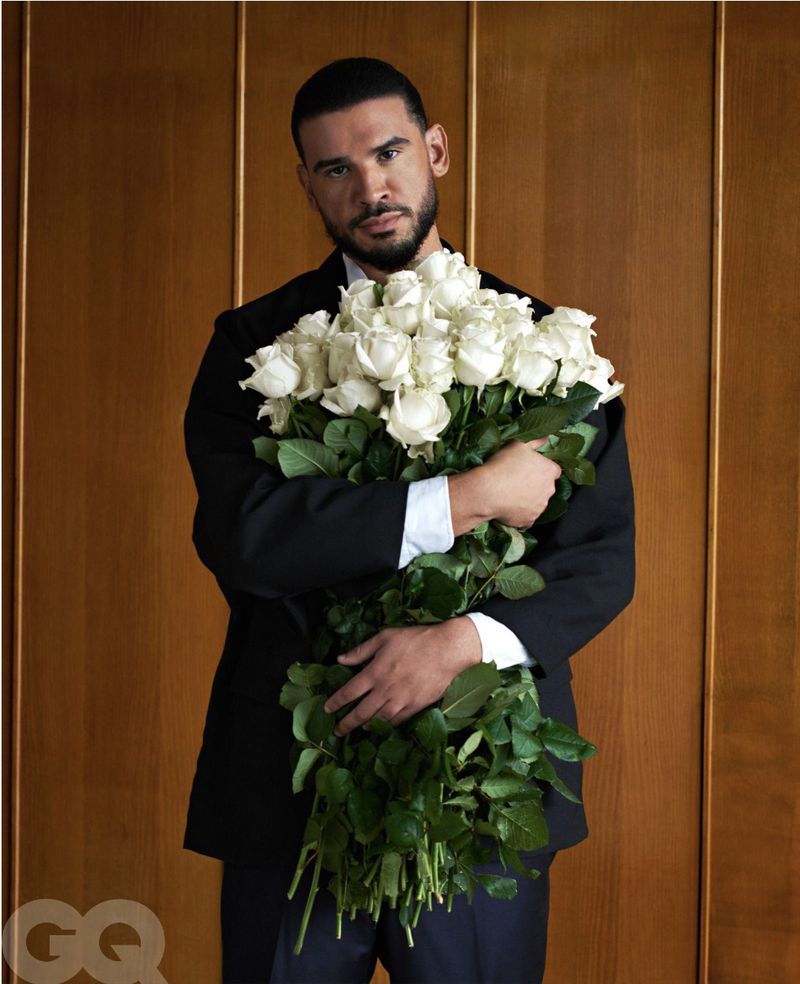 Adult male in a black suit holds a large bouquet of white roses against a wood-paneled background in a portrait shot.