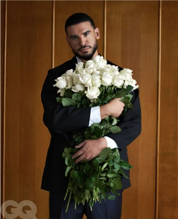 Adult male in a black suit holds a large bouquet of white roses against a wood-paneled background in a portrait shot.