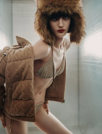 A model with red lipstick, wearing a trapper hat and bikini top, poses with a jacket slung over her shoulder in a photo studio.