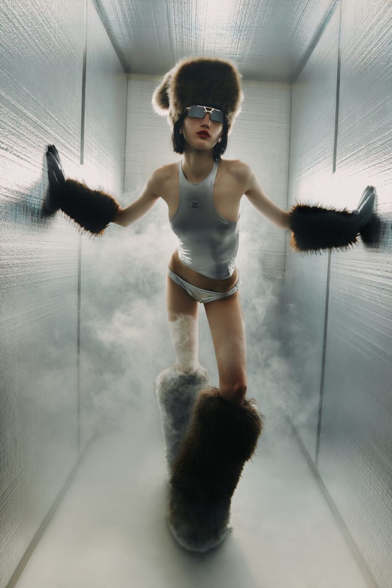 A woman in a silver swimsuit, fur hat, and fur arm and leg warmers poses in a minimalist, light-filled space with a smoky haze.