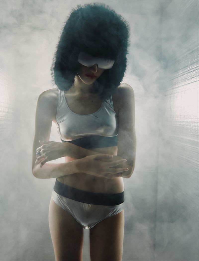 A woman in a white lingerie-style outfit with a large fur hat and goggles poses in a misty environment, arms crossed, looking down.