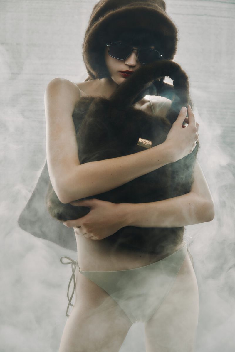 A woman in swimwear and sunglasses poses with a fur hat and handbag in a hazy photo studio setting for a fashion editorial.