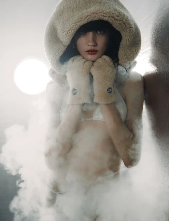 Young woman in hooded shearling coat and mittens poses indoors surrounded by fog with bright studio lighting, red lips, and bangs.