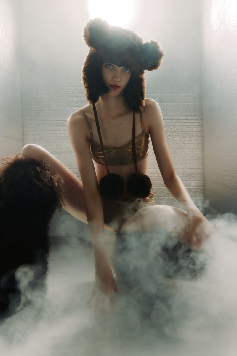 A woman in a bear hat and bikini poses in an indoor setting, surrounded by smoke or fog, creating a surreal and artistic fashion image.